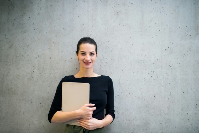 A PA holds a clipboard and stands against a concrete office wall.