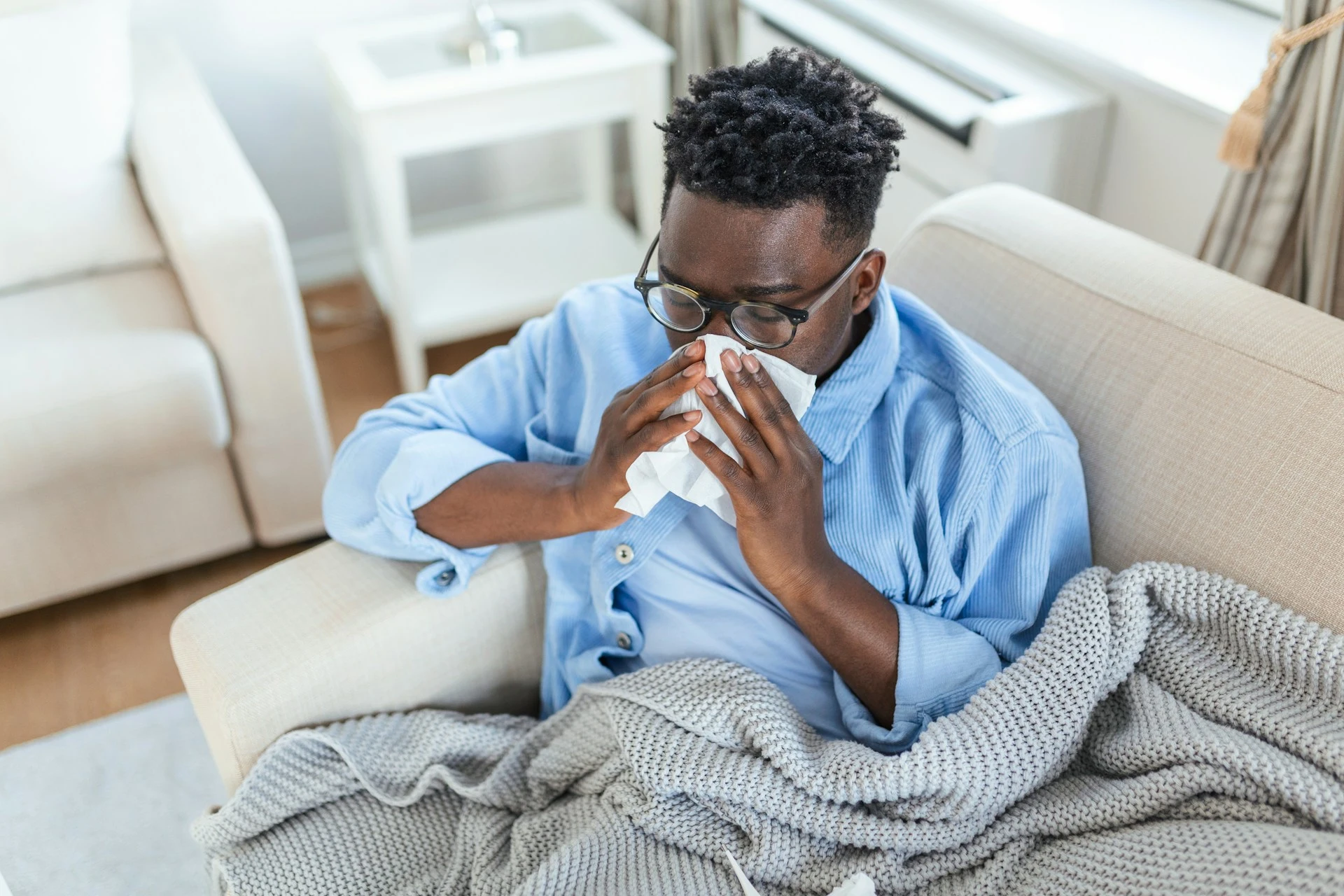 A professional man lays on the sofa in a blanket while blowing his nose. He is on long-term sick leave from work.