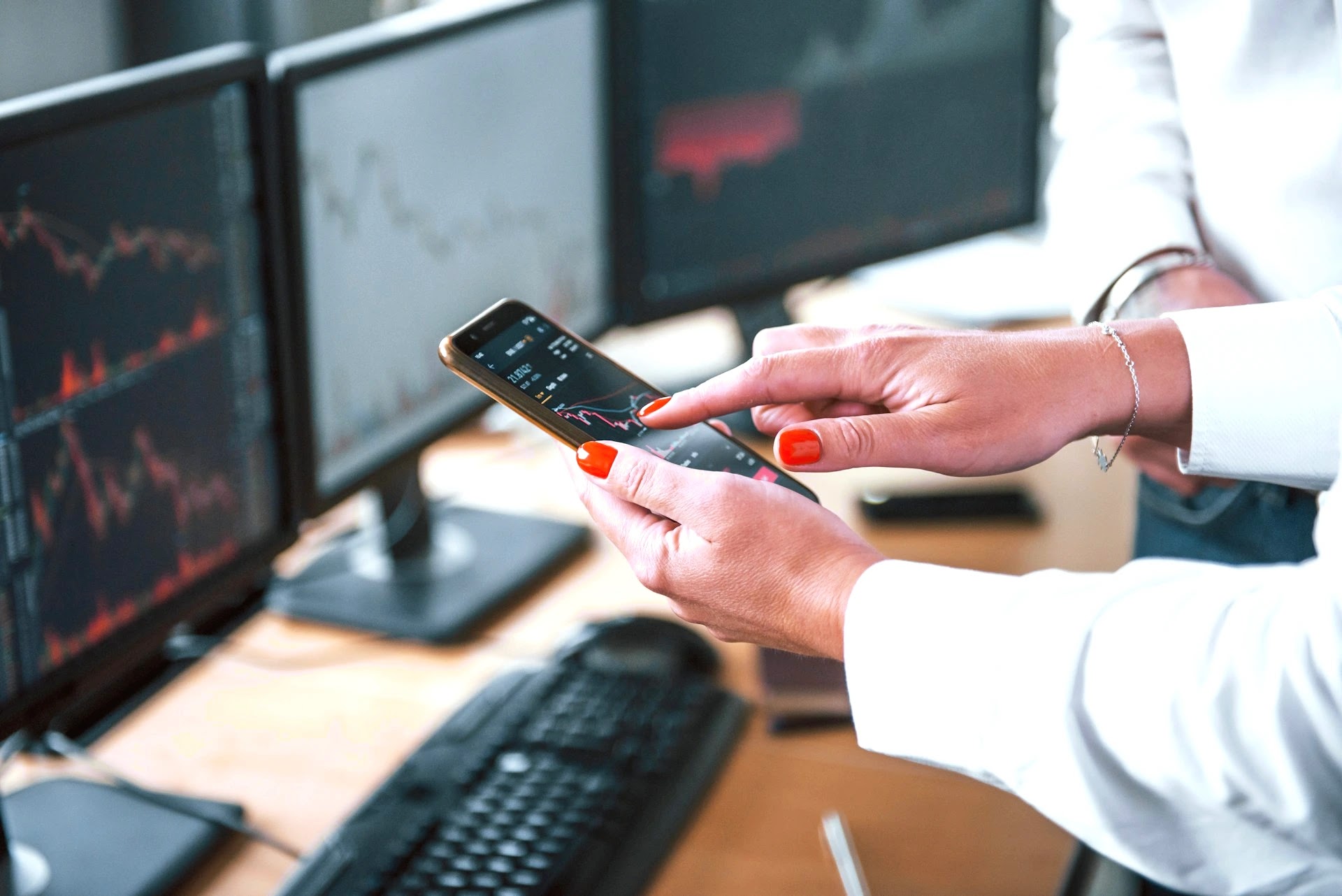 Close-up of hands checking financial market data on a smartphone in front of trading screens